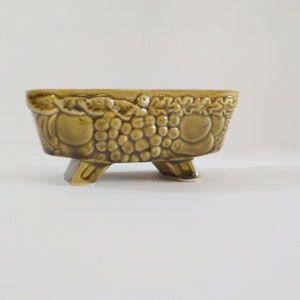 May include: A green ceramic planter with a raised design of grapes and apples. The planter has three legs and is oval shaped.
