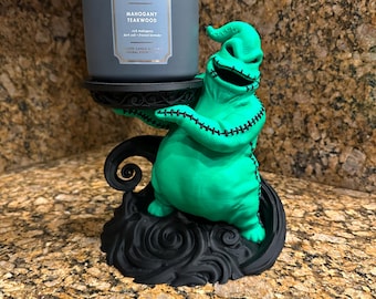 May include: A bright green, sack-like figure with black stitched details holds a black circular candle pedestal. A gray cylindrical candle jar labeled Mahogany Teakwood sits on the pedestal. The base is black with swirling patterns.
