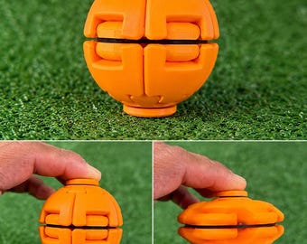 May include: A bright orange spherical fidget toy with a segmented, interlocking design. The object features a central black ring and can be compressed from a round ball shape into a flattened, disc-like form through its mechanical structure.