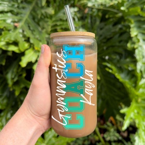 May include: A clear glass tumbler with a brown lid and a blue and green graphic that says "Coach Gymnastics". The tumbler is filled with a brown liquid and has a clear glass straw.