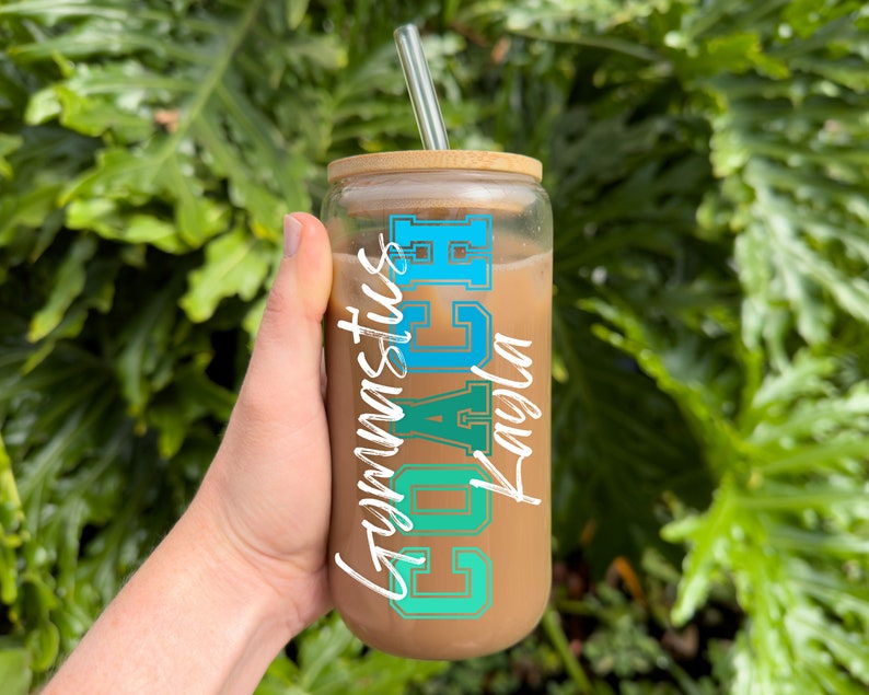 May include: A clear glass tumbler with a brown lid and a blue and green graphic that says "Coach Gymnastics". The tumbler is filled with a brown liquid and has a clear glass straw.