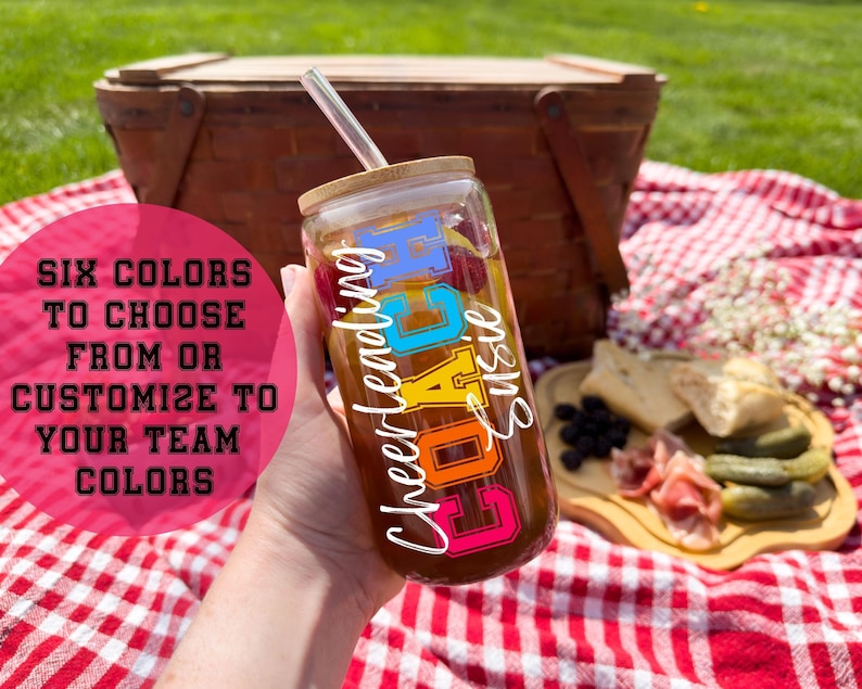 hand holding a glass cup with the words "Cheerleading COACH Susie" on it. Picnic blanket and basket in the background