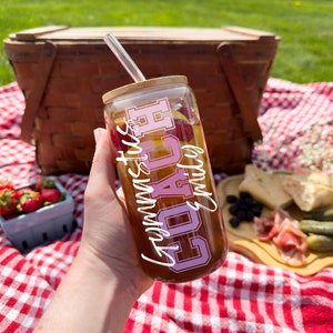 May include: A clear glass tumbler with a bamboo lid and a pink and white design that says "Gymnastics Coach" is being held by a hand. The tumbler is filled with a brown liquid and has a clear straw.