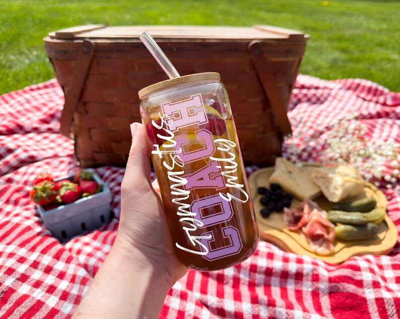 May include: A clear glass tumbler with a bamboo lid and a pink and white design that says "Gymnastics Coach" is being held by a hand. The tumbler is filled with a brown liquid and has a clear straw.