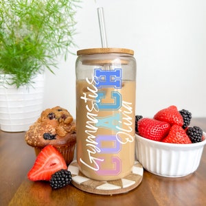 May include: A clear glass tumbler with a bamboo lid and straw. The tumbler is filled with iced coffee and has the text "Coach Olivia Gymnastics" written on it in colorful letters.