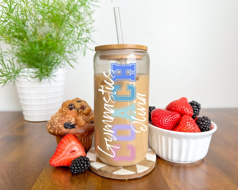 May include: A clear glass tumbler with a bamboo lid and straw. The tumbler is filled with iced coffee and has the text "Coach Olivia Gymnastics" written on it in colorful letters.