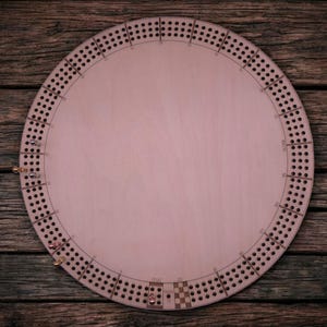 May include: A round wooden cribbage board with a light pink surface and a dark border. The board has numerous small holes for peg placement and is set against a rustic wooden background. Pegs are included.