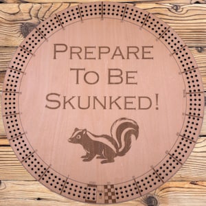 May include: Round wooden cribbage board with a skunk illustration and the text "PREPARE TO BE SKUNKED!". The board has holes for pegs and is designed for the classic card game. The board is made of light brown wood.
