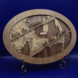 May include: A wooden oval-shaped sign with a map of Cape Cod and the text "Life is Better at the Cape". The sign is on a gold metal stand.