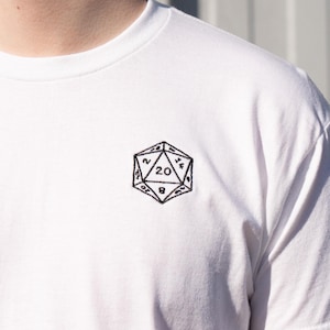 Peut inclure: T-shirt blanc avec un motif brodé noir d'un dé à 20 faces (D20) sur la poitrine. Le dé affiche le chiffre 20. Le t-shirt est fait d'une matière douce et légère, idéale pour un usage quotidien. Le design est simple et épuré.