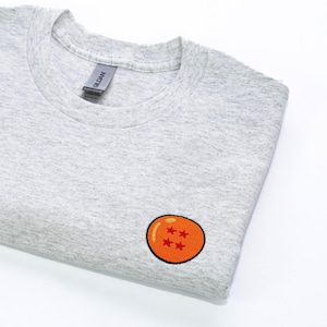 Op de afbeelding: Een gevouwen lichtgrijs sweatshirt met ronde hals. Het sweatshirt heeft een geborduurde oranje bol met vier rode sterren. Het Gildan-merklabel is zichtbaar bij de halslijn. Het shirt is gemaakt van een zacht, comfortabel materiaal.