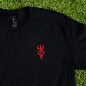May include: Black t-shirt with a red, stylized emblem on the chest. The emblem has a dripping effect, adding a unique visual element. The shirt is made of a soft material and is suitable for casual wear.
