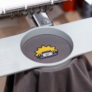 Op de afbeelding: Een grijs t-shirt met een geel en zwart geborduurd ontwerp van een hoofddeksel van een personage. Het shirt wordt op zijn plaats gehouden door een borduurmachine. Het ontwerp heeft een gele stekelige vorm met een grijs en wit embleem.