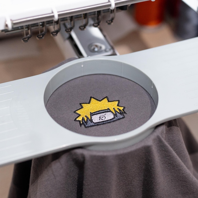Op de afbeelding: Een grijs t-shirt met een geel en zwart geborduurd ontwerp van een hoofddeksel van een personage. Het shirt wordt op zijn plaats gehouden door een borduurmachine. Het ontwerp heeft een gele stekelige vorm met een grijs en wit embleem.