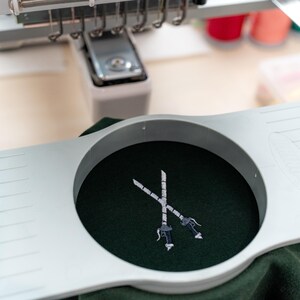 以下が含まれることがあります： 緑色の布地に、白い糸で刺繍された2本の交差した剣のデザインが施されています。布地は円形の刺繍枠に固定されています。