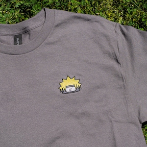 Op de afbeelding: Een grijs t-shirt met een klein geborduurd ontwerp van een geel en blauw anime-personage's hoofddeksel. Het t-shirt heeft een ronde hals en korte mouwen. Het Gildan-merklabel is zichtbaar op de kraag.