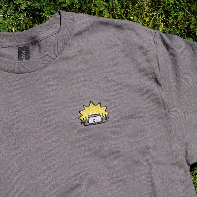 Op de afbeelding: Een grijs t-shirt met een klein geborduurd ontwerp van een geel en blauw anime-personage's hoofddeksel. Het t-shirt heeft een ronde hals en korte mouwen. Het Gildan-merklabel is zichtbaar op de kraag.