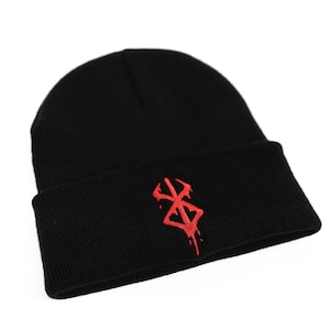 May include: Black knitted beanie hat featuring a red embroidered emblem on the front. The emblem is a stylized symbol with a dripping effect. The hat has a folded cuff and a rounded crown.
