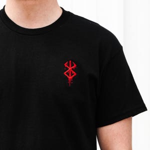 May include: Black crew neck t-shirt with a small, red, stylized emblem on the chest. The emblem has a symmetrical design with sharp angles and a dripping effect, suggesting a gothic or fantasy theme.