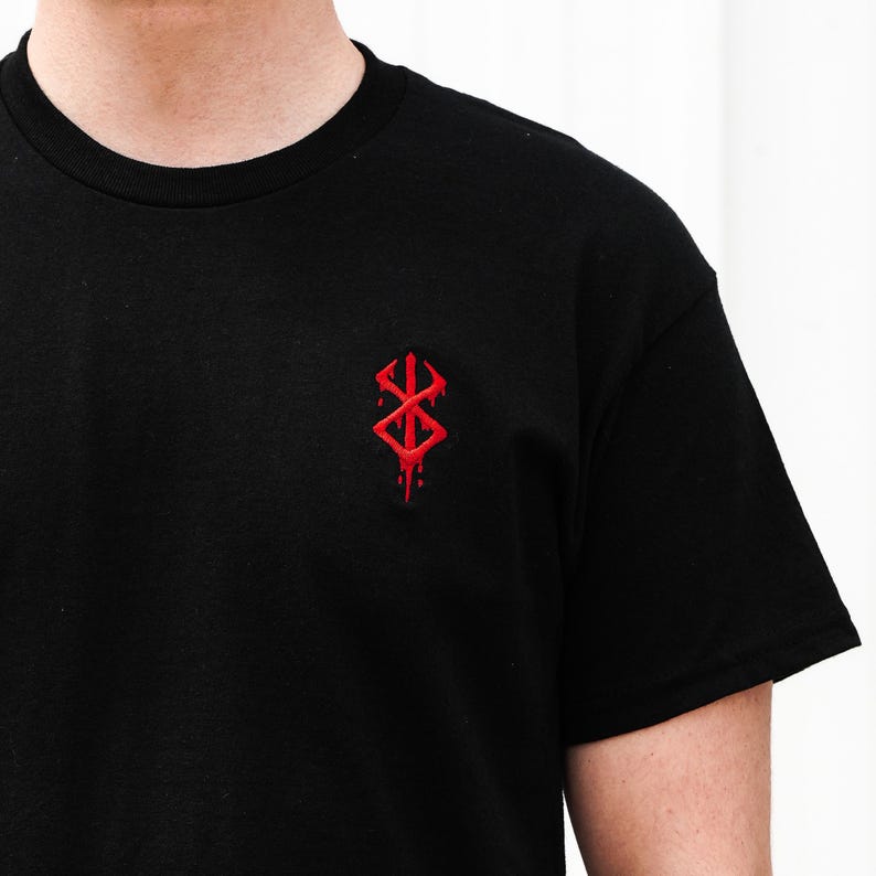 May include: Black crew neck t-shirt with a small, red, stylized emblem on the chest. The emblem has a symmetrical design with sharp angles and a dripping effect, suggesting a gothic or fantasy theme.