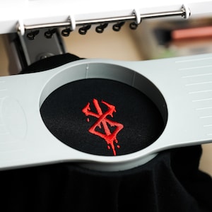 May include: Close-up of a black fabric with a red embroidered design. The fabric is held in place by a gray embroidery hoop. The design appears to be a stylized symbol.