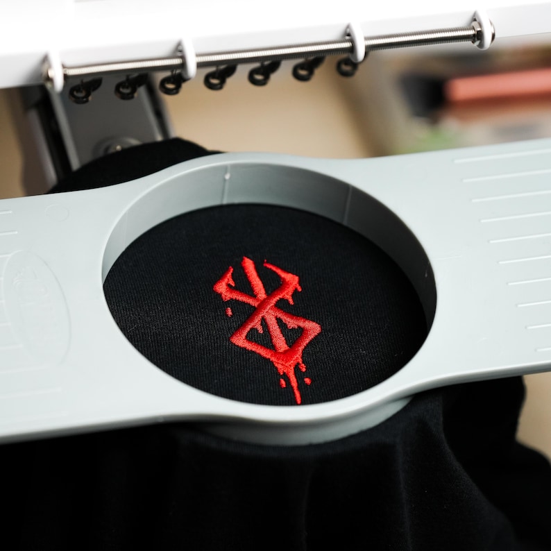 May include: Close-up of a black fabric with a red embroidered design. The fabric is held in place by a gray embroidery hoop. The design appears to be a stylized symbol.