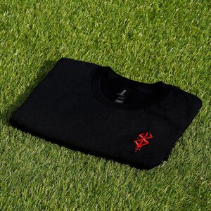 May include: Black crew neck sweatshirt with a red embroidered design on the chest. The sweatshirt is folded and resting on a bed of green grass. The Gildan brand tag is visible.