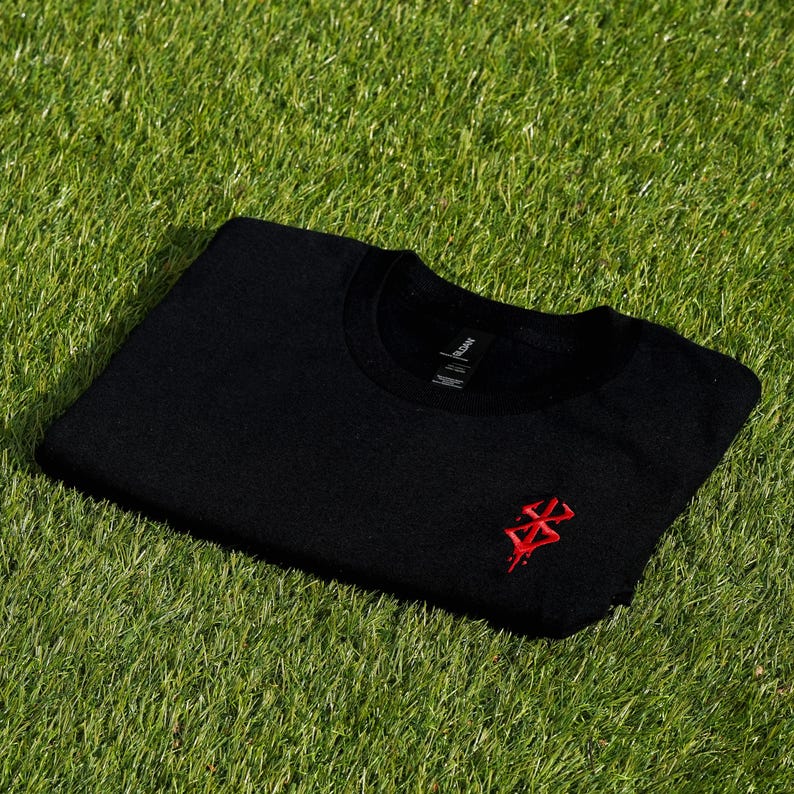 May include: Black crew neck sweatshirt with a red embroidered design on the chest. The sweatshirt is folded and resting on a bed of green grass. The Gildan brand tag is visible.