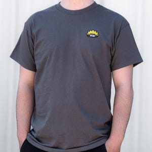 Op de afbeelding: Donkergrijs t-shirt met een klein, geborduurd ontwerp van een gele ster en een blauw-gele hoofdband. Het shirt heeft korte mouwen en een ronde hals.
