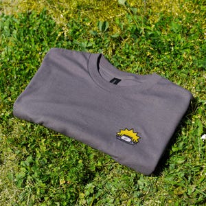 Op de afbeelding: Een gevouwen grijs crewneck T-shirt met een klein geborduurd anime-symbool in geel en zwart. Het T-shirt ligt op groen gras, met de kraag naar boven.