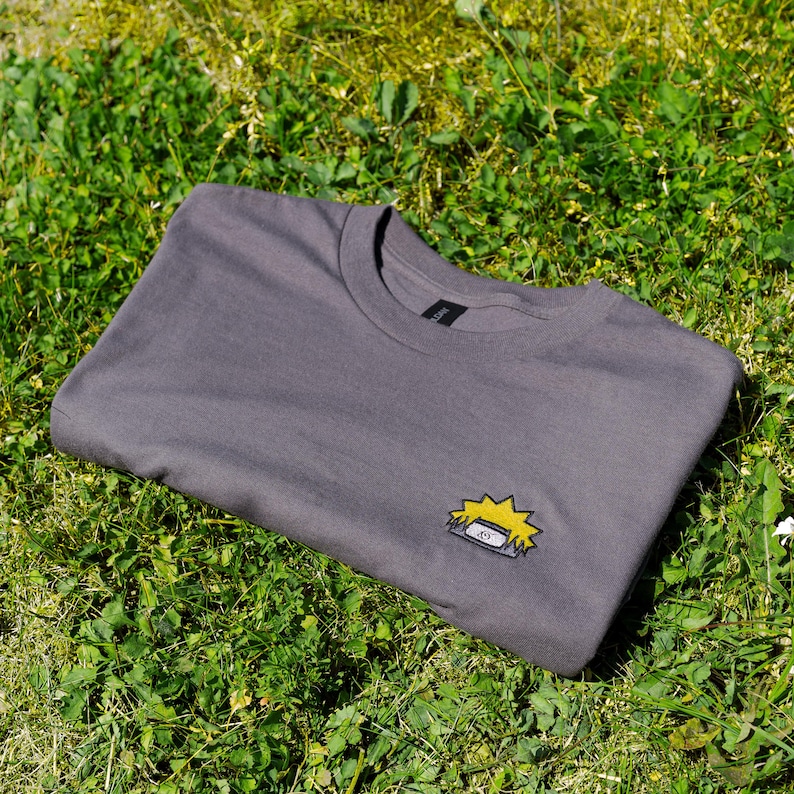 Op de afbeelding: Een gevouwen grijs crewneck T-shirt met een klein geborduurd anime-symbool in geel en zwart. Het T-shirt ligt op groen gras, met de kraag naar boven.