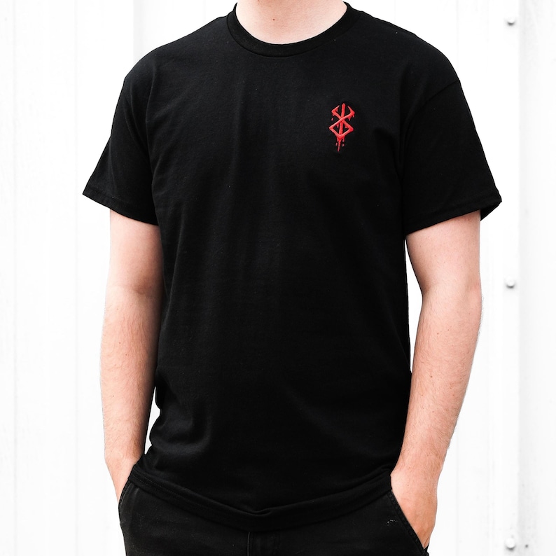 May include: Black t-shirt with a red graphic on the upper left chest. The graphic is a stylized symbol with a dripping effect. The shirt has short sleeves and a crew neck. The person is wearing black pants.