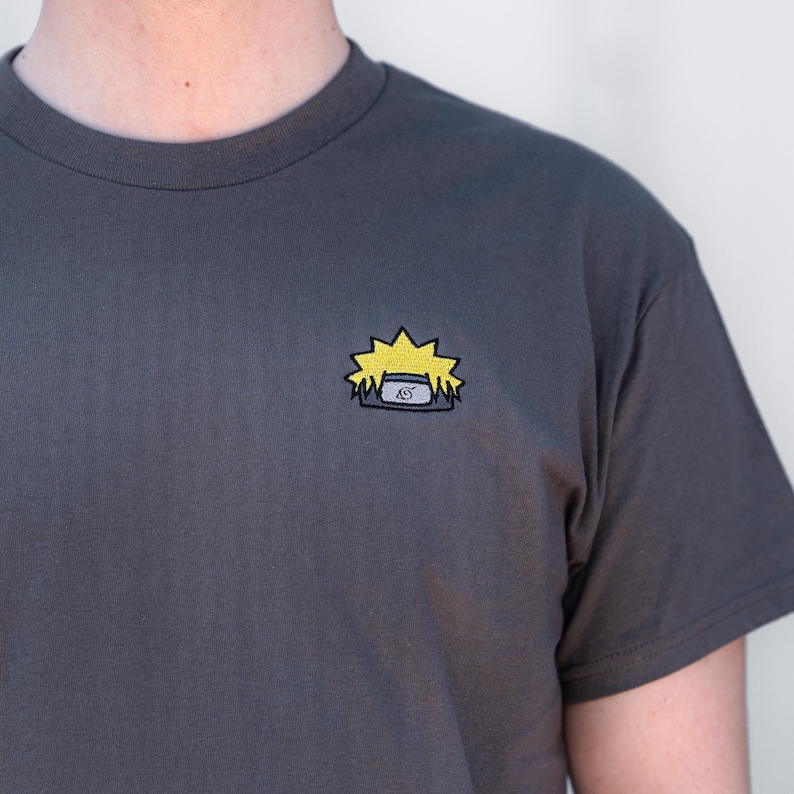 Op de afbeelding: Antracietgrijs T-shirt met een klein geborduurd ontwerp van gele stekelige haren en een blauw-wit symbool. Het shirt heeft korte mouwen en een ronde hals. Het ontwerp bevindt zich op de linkerborst.