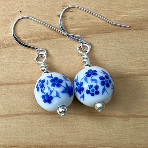 May include: A pair of dangle earrings featuring white ceramic beads with blue floral patterns. The beads are connected to silver-colored ear wires with a wrapped wire design. The earrings are displayed on a light brown wooden surface.