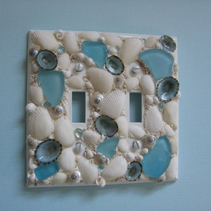 May include: A white light switch cover decorated with seashells and blue glass pieces.