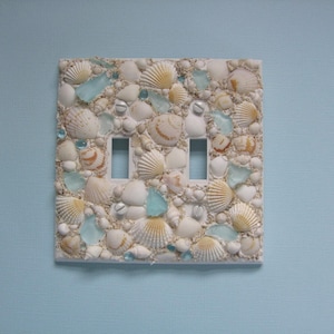 May include: A white light switch cover decorated with seashells and sea glass. The cover has three light switch openings.