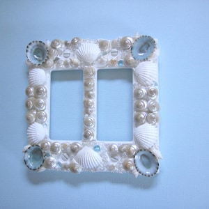 May include: A white double light switch cover decorated with seashells and iridescent beads. The cover has a light blue background and is surrounded by a variety of white and blue seashells.