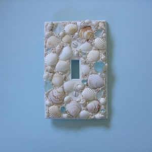 May include: A white light switch cover decorated with seashells and small pieces of blue glass.