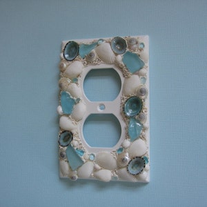May include: White outlet cover decorated with seashells and blue glass pieces.