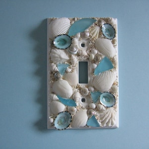 May include: White light switch cover decorated with seashells and blue glass pieces.