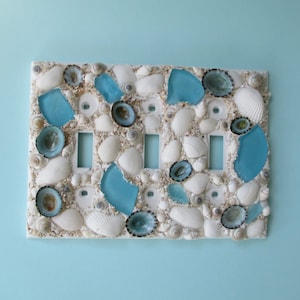 May include: A white light switch cover decorated with seashells and blue glass pieces.