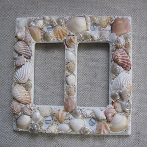 May include: White double light switch cover decorated with seashells in various shades of white, beige, and brown.