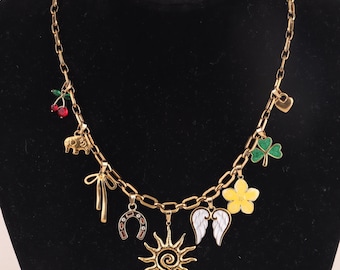 May include: A gold-toned chain necklace featuring multiple dangling charms including red cherries, an elephant, a ribbon bow, a horseshoe, a sun, white wings, a yellow flower, a green clover, and a small heart charm, all attached to the links.