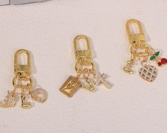 May include: Three gold-toned metal keychains with lobster clasps. The first features a narwhal, the letter L, and a crescent moon. The second has a rectangular tulip charm, the letter V, and a cross. The third includes the letter E, a heart, and cherries.