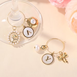 Könnte beinhalten: Goldfarbene Wein-Charms mit dekorativen Elementen. Zu den Charms gehören eine Blume, eine Schleife, eine Perle und Initialen. Die Charms sind an einem goldenen Ring befestigt, der zum Aufhängen an einem Weinglas bestimmt ist.