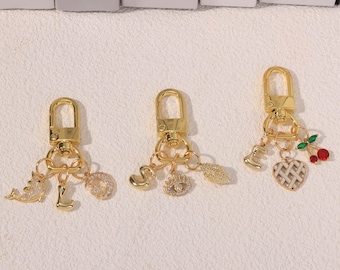 May include: Three gold-toned metal keychains with lobster clasps. The first features a dolphin, the letter L, and a crescent moon. The second has the letter S, an eye charm, and a leaf. The third includes the letter E, a heart, and cherries.