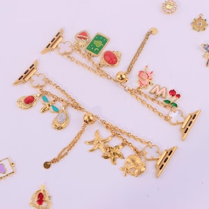 Könnte beinhalten: Goldfarbenes Uhrenarmband mit dekorativen Anhängern. Zu den Anhängern gehören ein roter Edelstein, eine grüne Karte, ein Schmetterling und ein Seestern. Das Armband wird an einer Smartwatch befestigt und hat ein Ketten-Design mit mehreren baumelnden Anhängern.