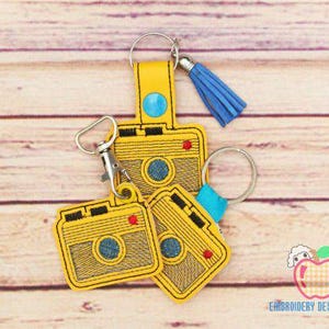 Könnte beinhalten: Drei gelbe, gestickte Schlüsselanhänger in Form einer Kamera mit blauen und roten Akzenten. Die Schlüsselanhänger sind an silbernen Schlüsselringen befestigt.