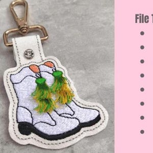 Puede incluir: Llavero de cuero blanco con un diseño bordado de dos botas blancas con borlas verdes y amarillas. El llavero tiene un anillo y un cierre de metal dorado. Tipos de archivo: DST, EXP, HUS, JEF, PES, VIP, VP3, XXX