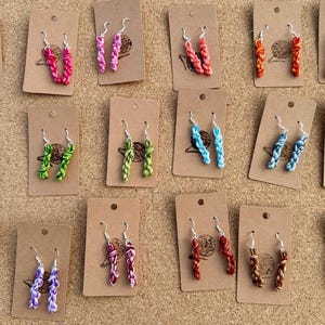 May include: A selection of braided rope earrings in a range of colours, such as red, pink, green, blue, orange, yellow, purple, and brown. Each pair is presented on a small, brown card with a silver hook.